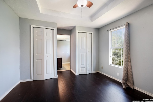 unfurnished bedroom with baseboards, a tray ceiling, multiple closets, and dark wood finished floors