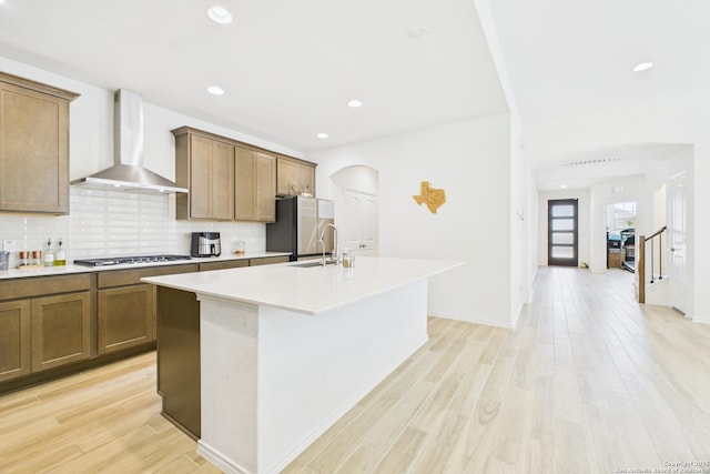 kitchen with wall chimney range hood, an island with sink, arched walkways, backsplash, and appliances with stainless steel finishes