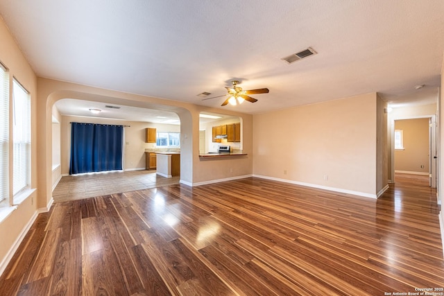 unfurnished living room featuring dark wood-style floors, a ceiling fan, visible vents, arched walkways, and plenty of natural light