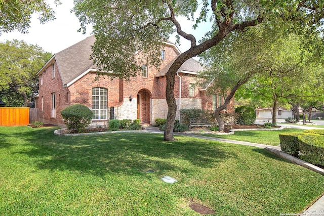tudor-style house featuring fence, brick siding, a front yard, and roof with shingles