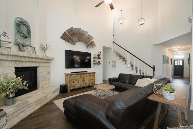 living room featuring a towering ceiling, stairs, ceiling fan, a fireplace, and dark wood-type flooring