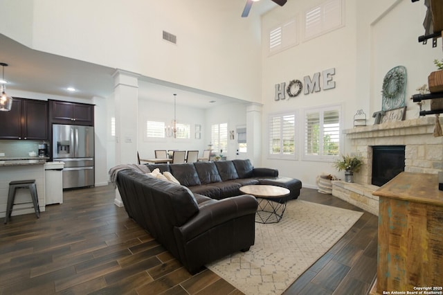 living area with a high ceiling, dark wood-style floors, visible vents, decorative columns, and ceiling fan with notable chandelier