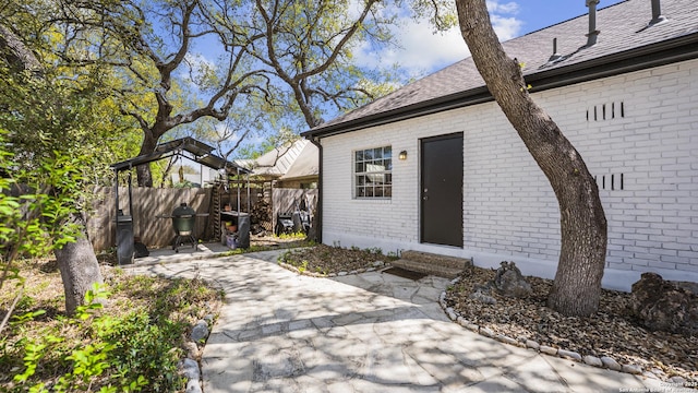 view of exterior entry featuring brick siding, fence, roof with shingles, and a patio