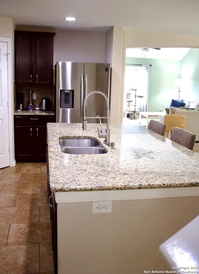 kitchen featuring light stone counters, stainless steel fridge with ice dispenser, dark brown cabinets, a sink, and a center island with sink