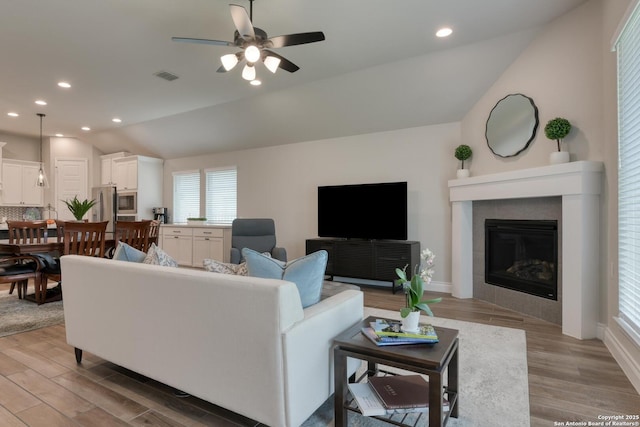 living room with ceiling fan, a tile fireplace, light wood finished floors, lofted ceiling, and visible vents