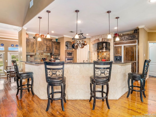 kitchen featuring built in appliances, light wood-type flooring, wall chimney exhaust hood, and crown molding
