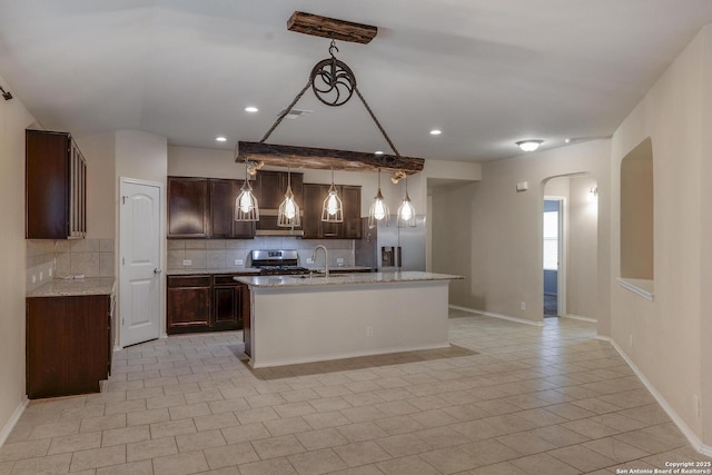 kitchen with stainless steel appliances, dark brown cabinets, arched walkways, a kitchen island with sink, and backsplash