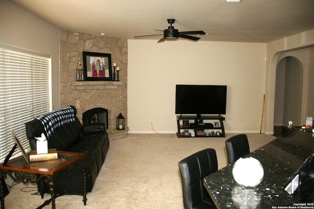 living room with arched walkways, a stone fireplace, carpet flooring, baseboards, and a ceiling fan
