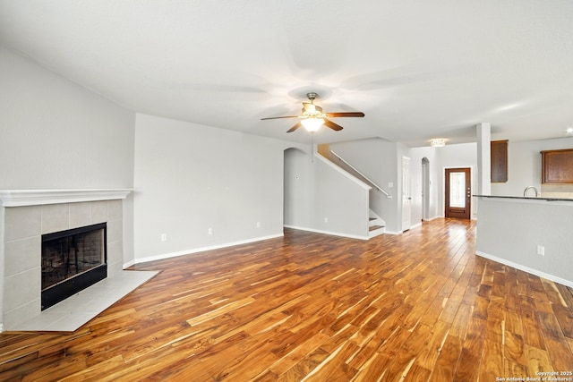 unfurnished living room with stairway, ceiling fan, arched walkways, wood finished floors, and a tiled fireplace