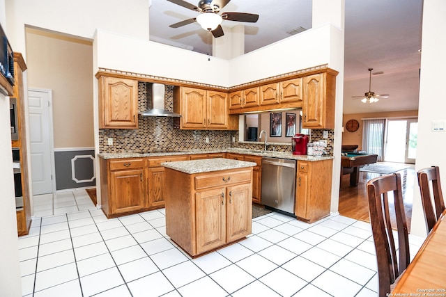 kitchen featuring wall chimney range hood, stainless steel dishwasher, a kitchen island, light tile patterned flooring, and a ceiling fan