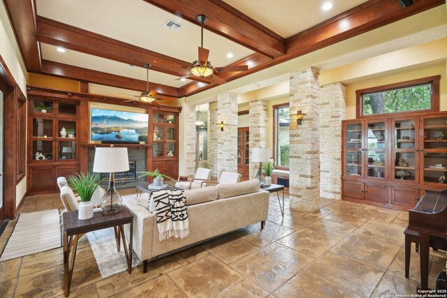 living area featuring beamed ceiling, visible vents, decorative columns, a fireplace, and a ceiling fan