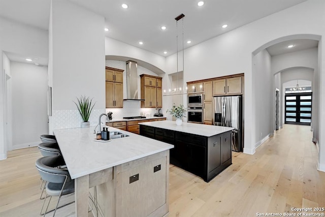 kitchen with light wood-style floors, appliances with stainless steel finishes, a sink, a center island, and wall chimney exhaust hood
