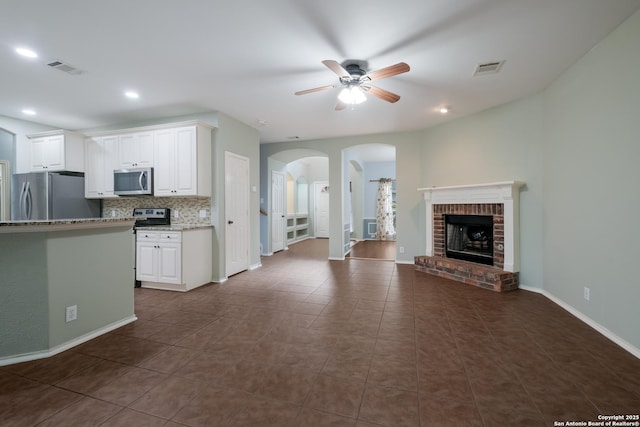 unfurnished living room with ceiling fan, baseboards, arched walkways, a brick fireplace, and dark tile patterned flooring