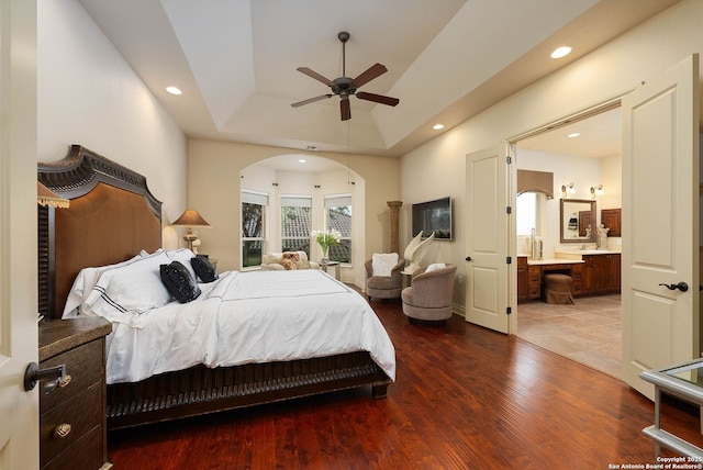 bedroom with wood finished floors, a raised ceiling, ensuite bath, and arched walkways