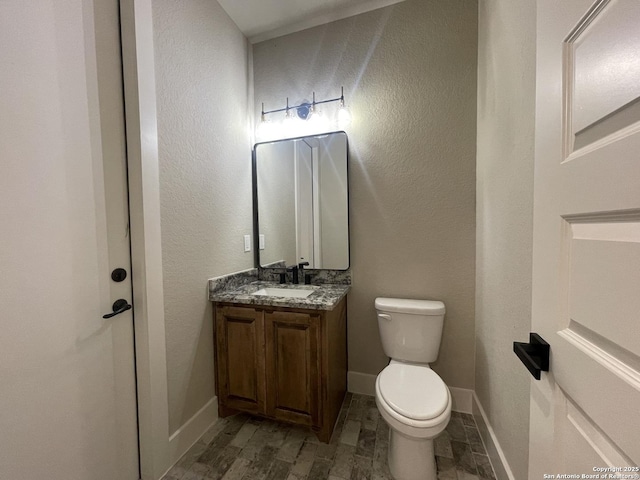 half bath featuring vanity, toilet, a textured wall, and baseboards