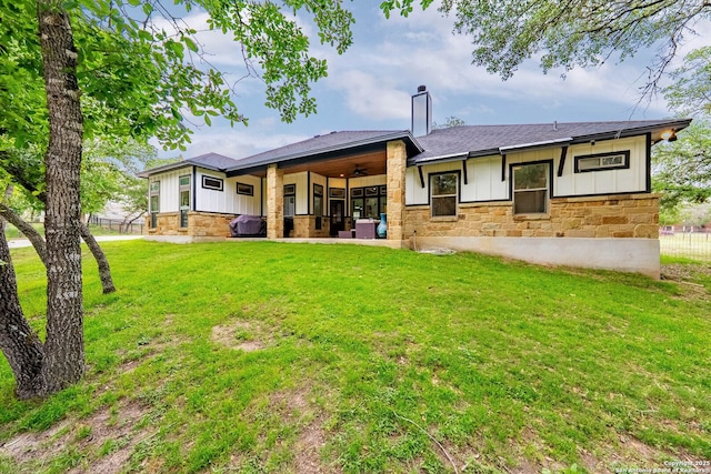 back of property with board and batten siding, a ceiling fan, a yard, and stone siding