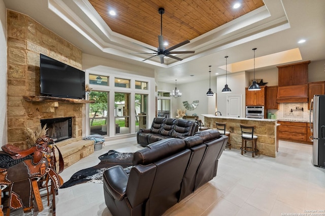 living area with a tray ceiling, wooden ceiling, a fireplace, a towering ceiling, and ceiling fan with notable chandelier