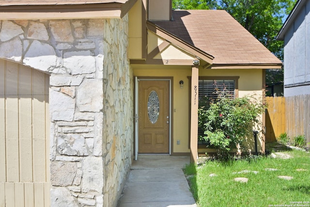 view of exterior entry featuring stone siding, stucco siding, a shingled roof, and fence