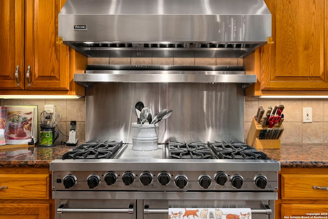 kitchen featuring double oven range, dark stone counters, decorative backsplash, brown cabinetry, and wall chimney range hood