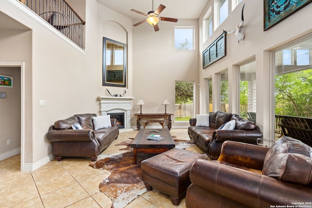 living area featuring light tile patterned floors, a lit fireplace, baseboards, a ceiling fan, and a high ceiling
