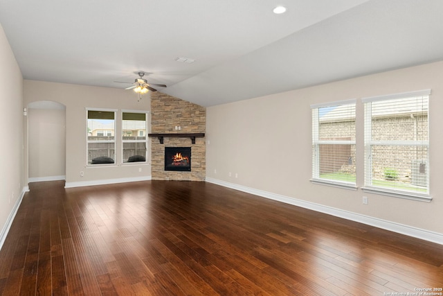 unfurnished living room featuring ceiling fan, arched walkways, a stone fireplace, vaulted ceiling, and dark wood-style floors