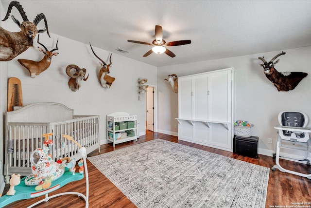 bedroom featuring ceiling fan, visible vents, a nursery area, baseboards, and wood finished floors