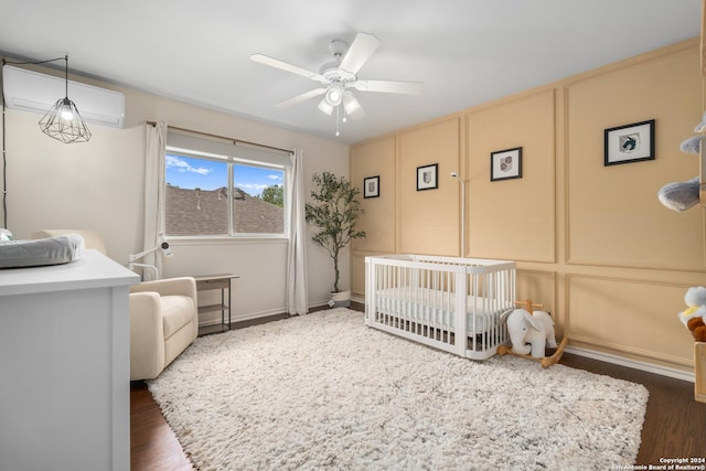 bedroom featuring ceiling fan, dark wood-style flooring, a wall mounted air conditioner, a crib, and a decorative wall