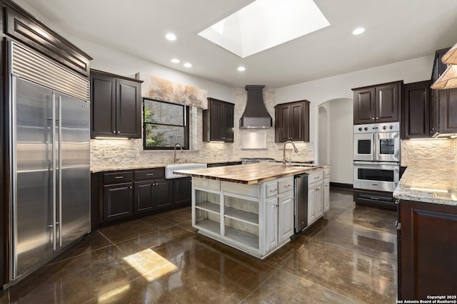 kitchen featuring appliances with stainless steel finishes, a sink, wooden counters, premium range hood, and decorative backsplash