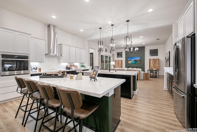 kitchen featuring an island with sink, wall chimney exhaust hood, a sink, light wood finished floors, and stainless steel appliances