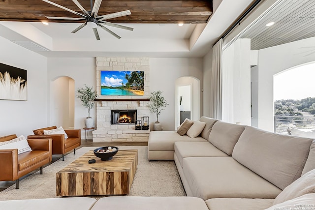 living area with a ceiling fan, arched walkways, a fireplace, a raised ceiling, and light wood finished floors