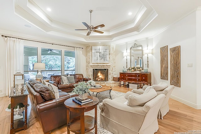 living room featuring crown molding, light wood-style floors, a stone fireplace, ceiling fan, and a tray ceiling