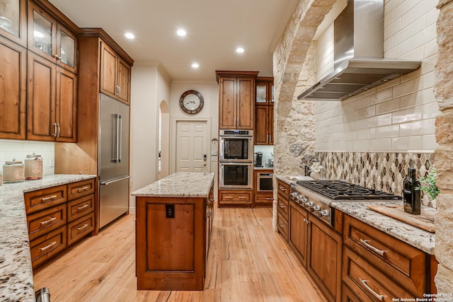 kitchen with wall chimney range hood, appliances with stainless steel finishes, light wood-style floors, a center island, and ornamental molding