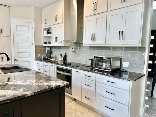 kitchen featuring oven, black electric stovetop, wall chimney range hood, dark stone counters, and white cabinetry