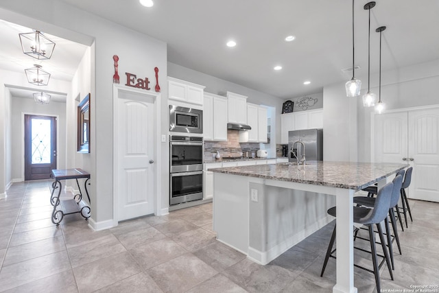 kitchen with light stone countertops, an island with sink, stainless steel appliances, backsplash, and under cabinet range hood