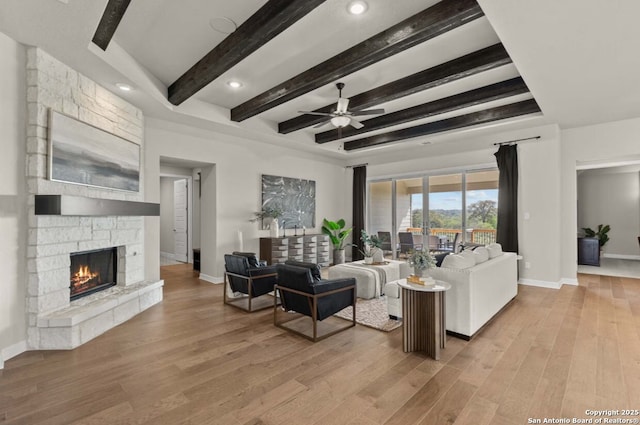 living room featuring a ceiling fan, a stone fireplace, baseboards, beamed ceiling, and light wood finished floors