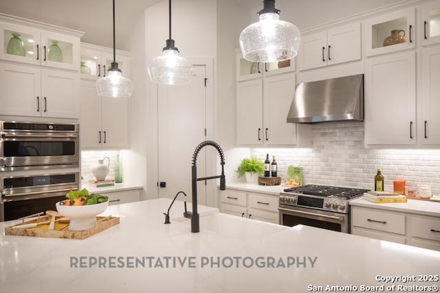 kitchen with wall chimney exhaust hood, white cabinets, appliances with stainless steel finishes, backsplash, and decorative light fixtures