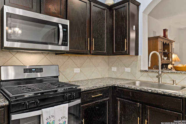 kitchen featuring dark brown cabinetry, backsplash, appliances with stainless steel finishes, arched walkways, and a sink