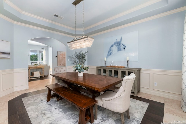 dining space with crown molding, a decorative wall, a wainscoted wall, a tray ceiling, and arched walkways