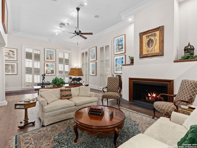 living area featuring a ceiling fan, wood finished floors, baseboards, a warm lit fireplace, and ornamental molding