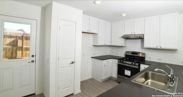 kitchen with dark countertops, a sink, stainless steel gas range oven, light wood-style flooring, and under cabinet range hood