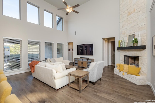 living room with a ceiling fan, a stone fireplace, a towering ceiling, wood finished floors, and baseboards