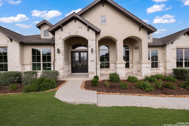 doorway to property featuring french doors, a lawn, a shingled roof, stone siding, and stucco siding