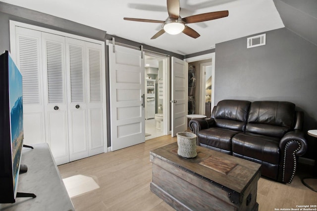living room with ceiling fan, stacked washer / dryer, visible vents, a barn door, and light wood-style floors