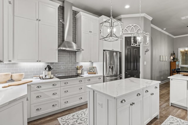 kitchen with wall chimney range hood, crown molding, white cabinets, stainless steel fridge, and a chandelier