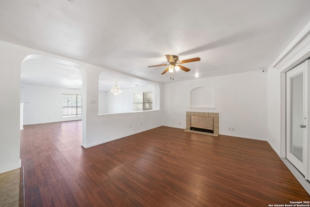 unfurnished living room featuring baseboards, dark wood-style floors, a tile fireplace, arched walkways, and ceiling fan with notable chandelier