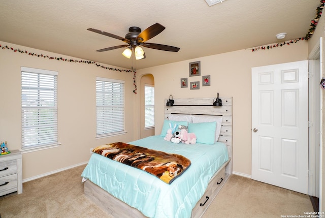 bedroom featuring light colored carpet, arched walkways, a textured ceiling, a ceiling fan, and baseboards