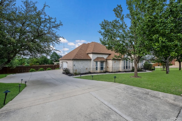 french country inspired facade with a garage, a front lawn, fence, stone siding, and concrete driveway