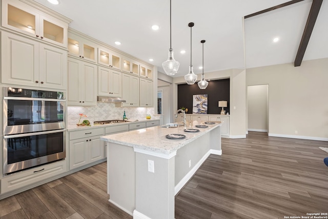 kitchen with wood finished floors, a kitchen island with sink, double oven, a sink, and backsplash