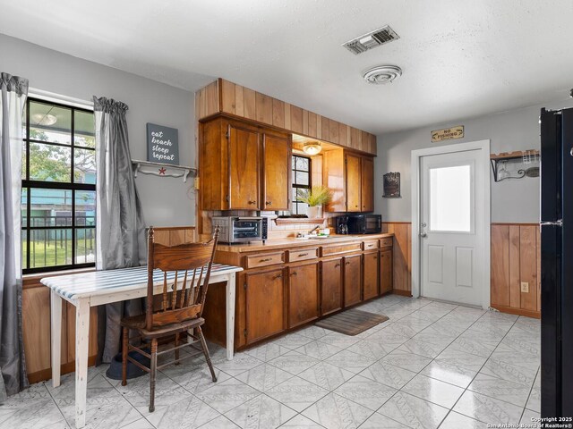 kitchen featuring brown cabinetry, visible vents, black microwave, light countertops, and a wainscoted wall