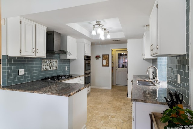 kitchen featuring appliances with stainless steel finishes, a sink, wall chimney exhaust hood, dark stone countertops, and a tray ceiling